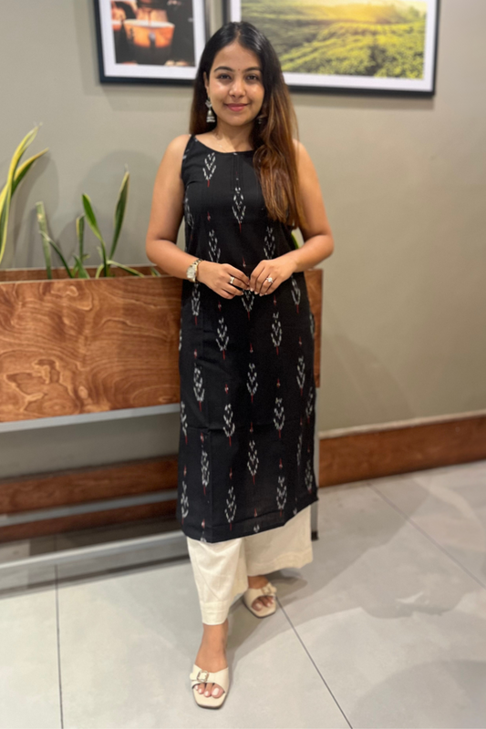Woman wearing a black patterned dress standing indoors with plants and framed pictures in the background.